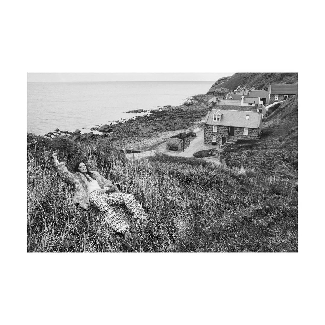 Black and white photo of a woman lying in tall grass by the sea, with stone houses on a hill in the background, creating a vintage aesthetic.