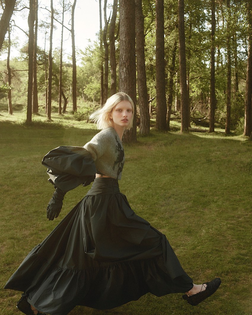 A woman in a long black skirt and fuzzy sweater poses in a forest setting, exhibiting high fashion and natural light. The woman is walking.