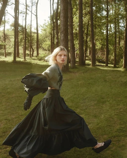 A woman in a long black skirt and fuzzy sweater poses in a forest setting, exhibiting high fashion and natural light. The woman is walking.