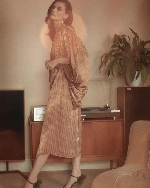 A woman in a gold, draped dress and heels poses indoors with vintage audio equipment, houseplants, and soft, diffused light.