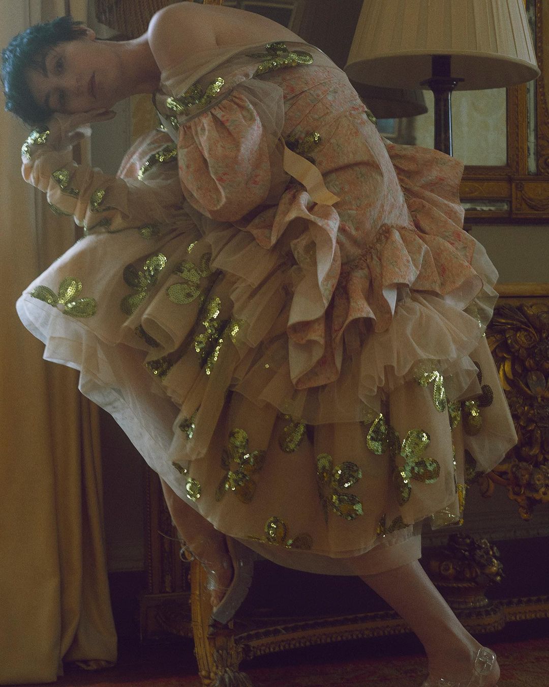 Elegant woman in a floral dress and gold sequins. She is indoors and posing next to a gold ornate table, with a warm colour grade.