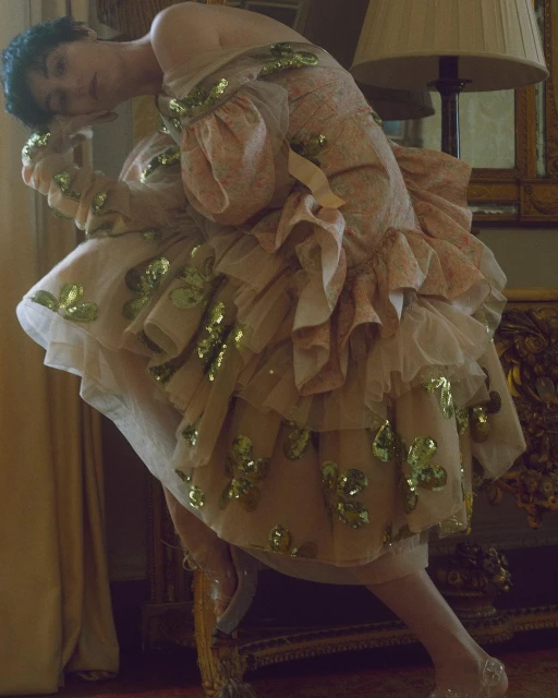 Elegant woman in a floral dress and gold sequins. She is indoors and posing next to a gold ornate table, with a warm colour grade.