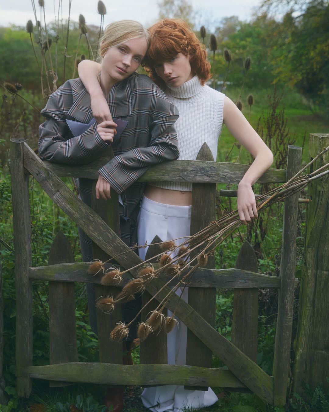 Two young women in fashionable outfits lean on a wooden gate surrounded by foliage in a daytime outdoor fashion editorial shot.