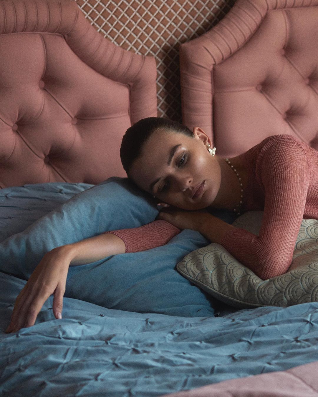 Woman rests on a bed with light blue bedding and headboard. She wears a peach top, pearl necklace, and flower earrings in an indoor bedroom setting.