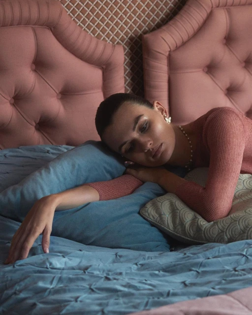 Woman rests on a bed with light blue bedding and headboard. She wears a peach top, pearl necklace, and flower earrings in an indoor bedroom setting.