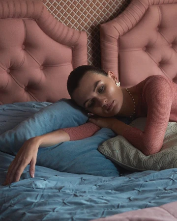 Woman rests on a bed with light blue bedding and headboard. She wears a peach top, pearl necklace, and flower earrings in an indoor bedroom setting.
