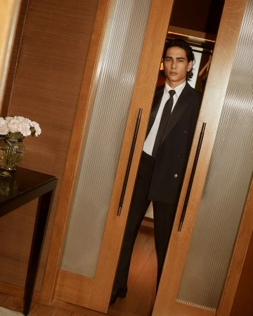 Young adult male poses confidently in a double-breasted black suit between frosted-glass paneled doors, showcasing fashion and style.