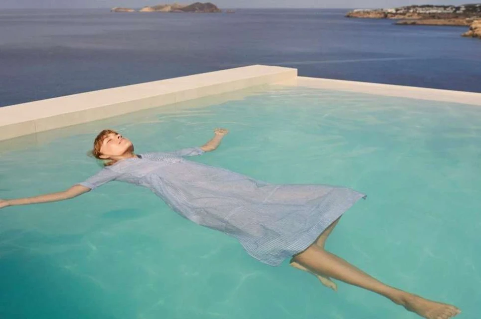 Woman in a dress floating in a blue swimming pool with ocean and islands in the background on a sunny day. Relaxation and tranquility.