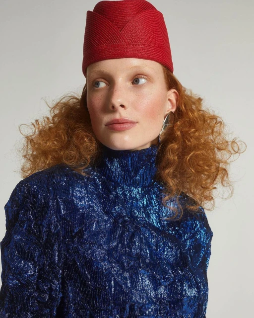 Red-haired woman with red hat and blue shiny textured top poses in a studio setting against a neutral background. Fashion editorial.