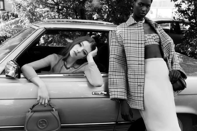 Two female models posing with a vintage car, one seated inside and leaning on the window, the other standing beside the vehicle outdoors.