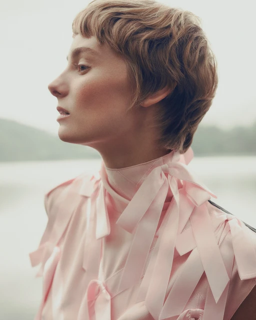 Profile shot of an adult female with blonde, wavy hair in a pink blouse with bow details, posed outdoors with a blurred waterscape background.