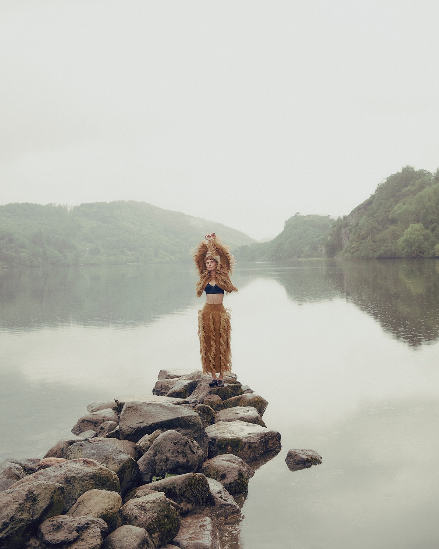 A woman stands on rocks in a lake, wearing a brown furry outfit, with misty mountains and water reflecting the sky in the background.