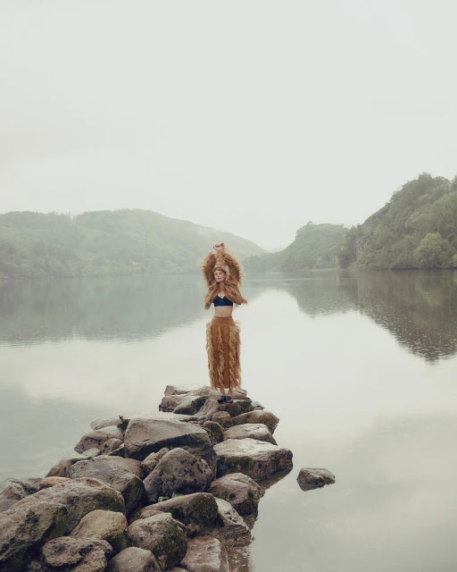 A woman stands on rocks in a lake, wearing a brown furry outfit, with misty mountains and water reflecting the sky in the background.