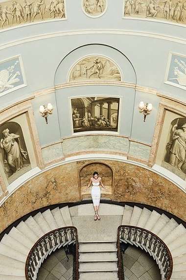 Overhead shot of a woman in a white dress lying on a stairway, surrounded by architectural details and classical-style artwork.