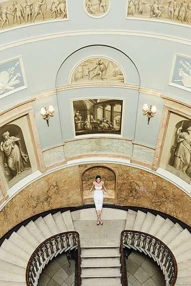 Overhead shot of a woman in a white dress lying on a stairway, surrounded by architectural details and classical-style artwork.