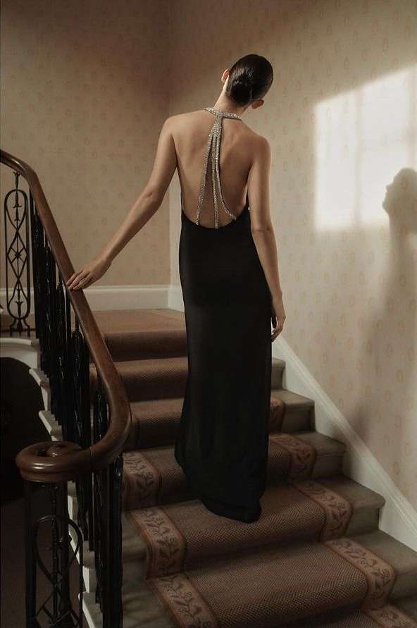 A woman in a black dress ascends carpeted stairs, back to the camera, her beaded halter adds detail to a neutral-toned interior.