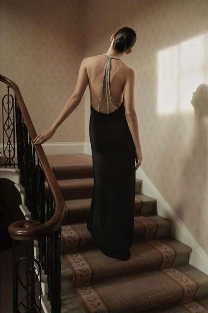 A woman in a black dress ascends carpeted stairs, back to the camera, her beaded halter adds detail to a neutral-toned interior.