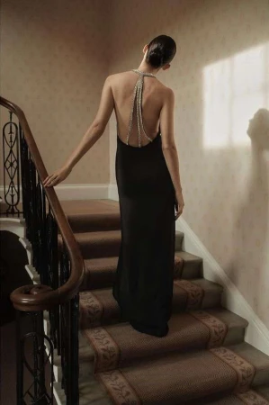 A woman in a black dress ascends carpeted stairs, back to the camera, her beaded halter adds detail to a neutral-toned interior.