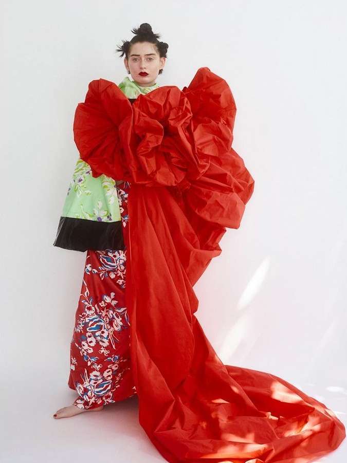 Fashion portrait of a woman in a bold red flower-like garment with floral pants, posing in a studio setting against a bright white backdrop.