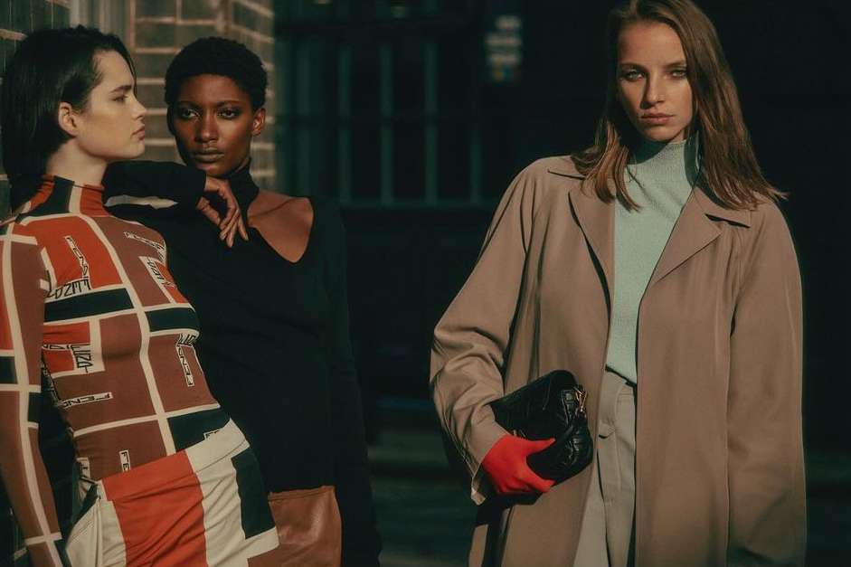 Three diverse women stand near a brick wall in fashionable outfits, bathed in natural light for a striking fashion editorial shot.