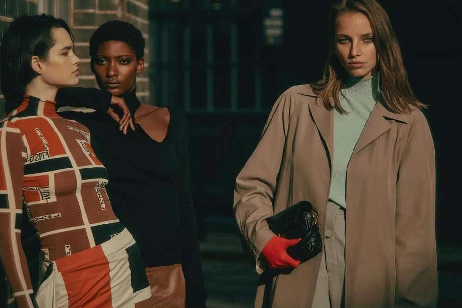 Three diverse women stand near a brick wall in fashionable outfits, bathed in natural light for a striking fashion editorial shot.