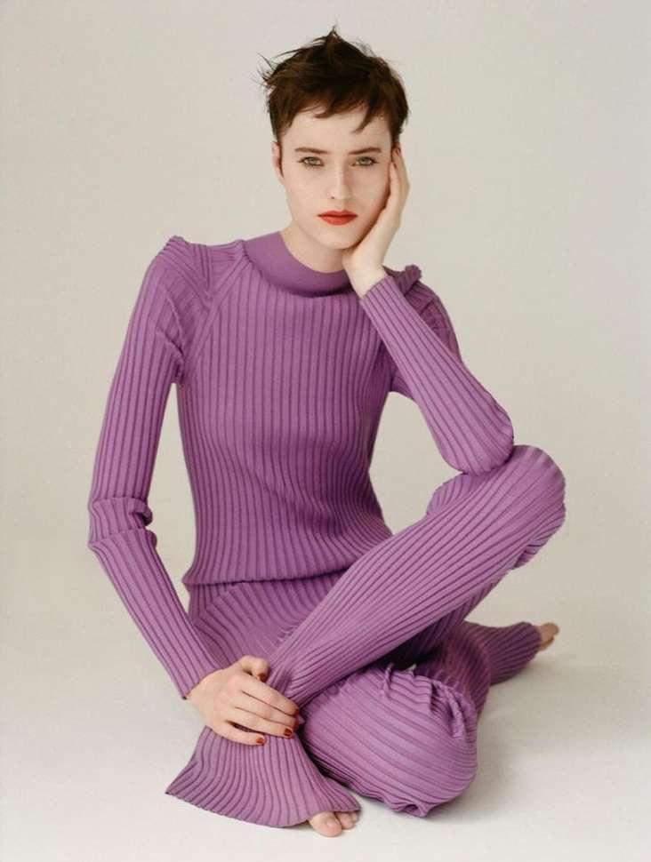 Fashion portrait of a woman with short brown hair, sitting cross-legged in a ribbed purple sweater and pants, against a neutral backdrop.
