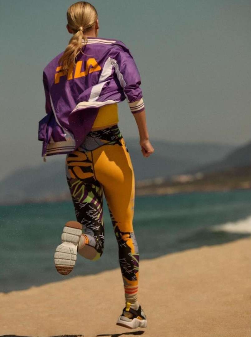 Model with blonde hair running along a sandy beach wearing a purple FILA jacket and patterned yellow leggings.