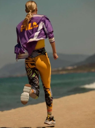 Model with blonde hair running along a sandy beach wearing a purple FILA jacket and patterned yellow leggings.
