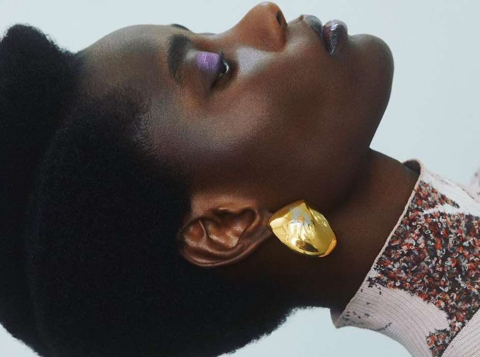 Close-up portrait of a black female with afro, gold earring, light purple eye shadow, glossy lips, and sequined fabric against a white background.