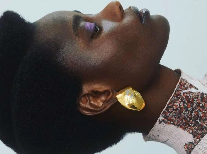 Close-up portrait of a black female with afro, gold earring, light purple eye shadow, glossy lips, and sequined fabric against a white background.