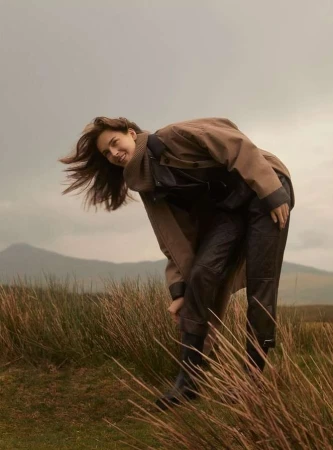Woman in brown coat and dark trousers leaning over in a grassy field with hills under an overcast sky by Agata Pospieszynska
