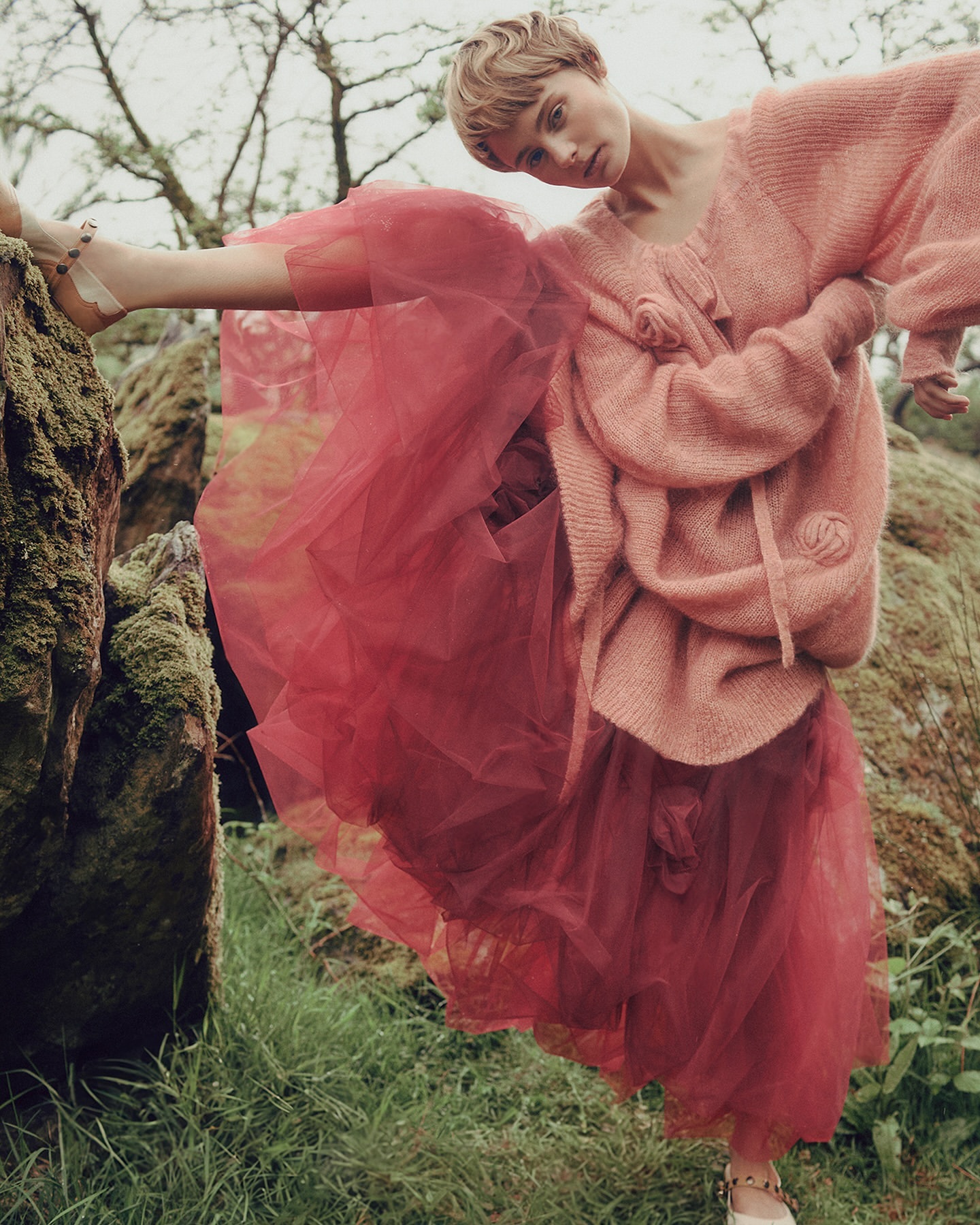 Fashion editorial shot of a young adult woman in a pink sweater and red tulle skirt, striking a pose in a mossy forest environment.