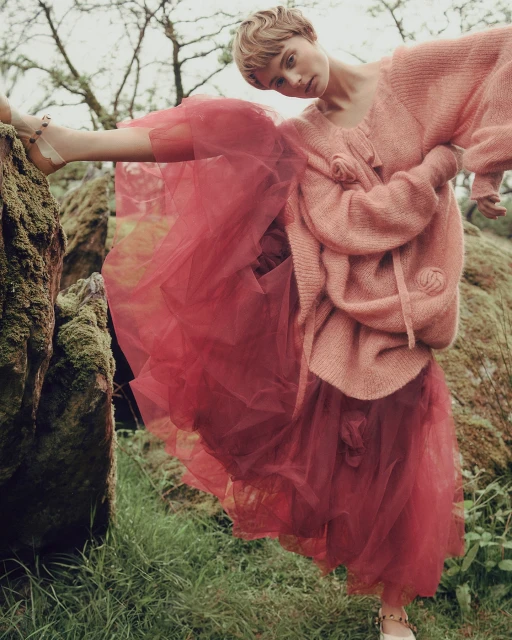 Fashion editorial shot of a young adult woman in a pink sweater and red tulle skirt, striking a pose in a mossy forest environment.
