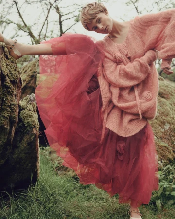 Fashion editorial shot of a young adult woman in a pink sweater and red tulle skirt, striking a pose in a mossy forest environment.