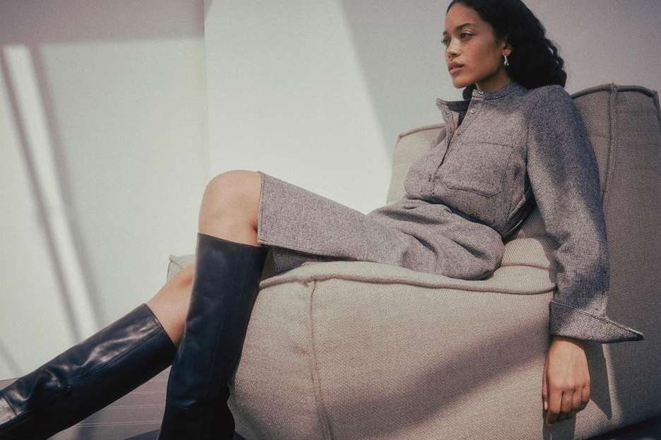 A woman in a gray dress and black boots sits on a couch in front of a white wall, illuminated by natural light.