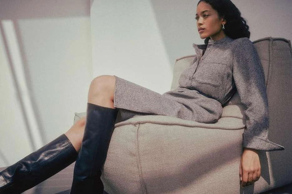 A woman in a gray dress and black boots sits on a couch in front of a white wall, illuminated by natural light.