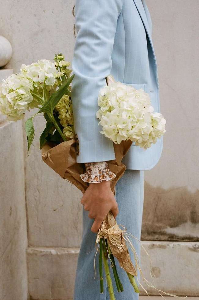 Person in a light blue suit holding a bouquet of white hydrangeas wrapped in brown paper, standing against a neutral wall. 