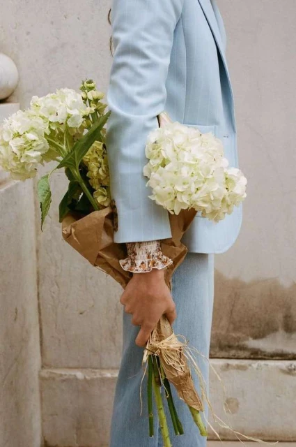 Person in a light blue suit holding a bouquet of white hydrangeas wrapped in brown paper, standing against a neutral wall. 