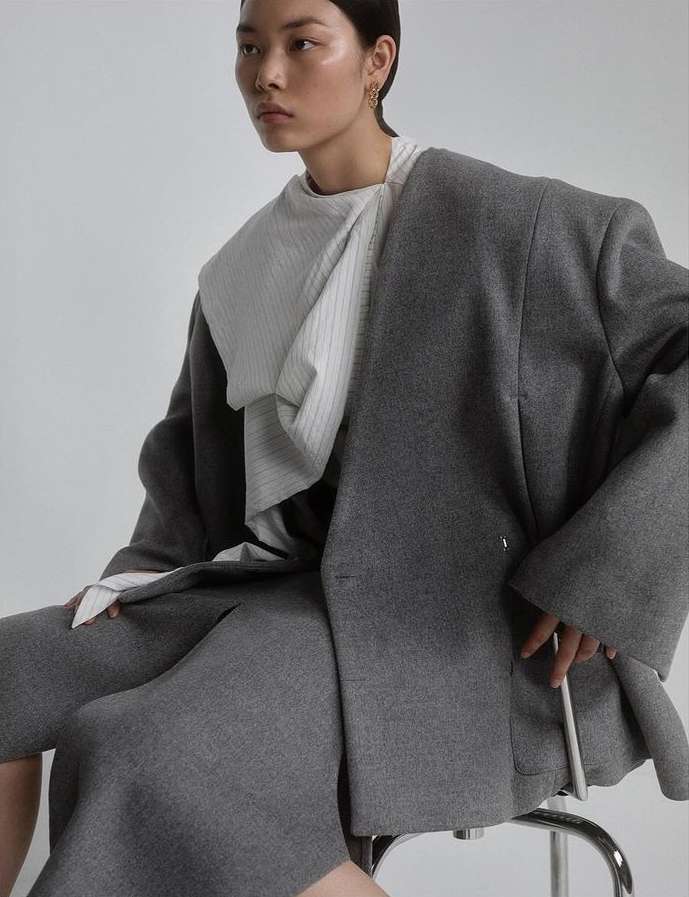 Asian fashion model poses in grey wool blazer and skirt, seated on a silver stool in a bright studio, exuding minimalist style.