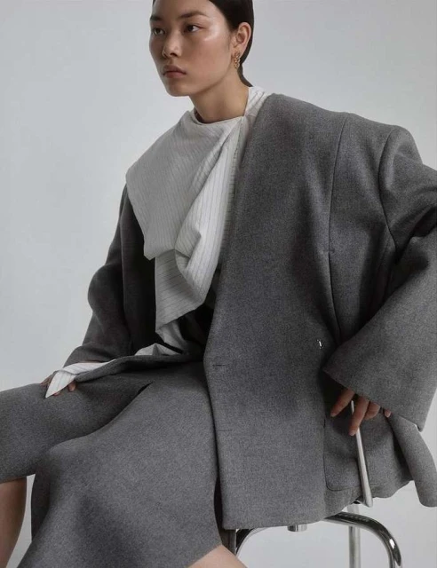 Asian fashion model poses in grey wool blazer and skirt, seated on a silver stool in a bright studio, exuding minimalist style.
