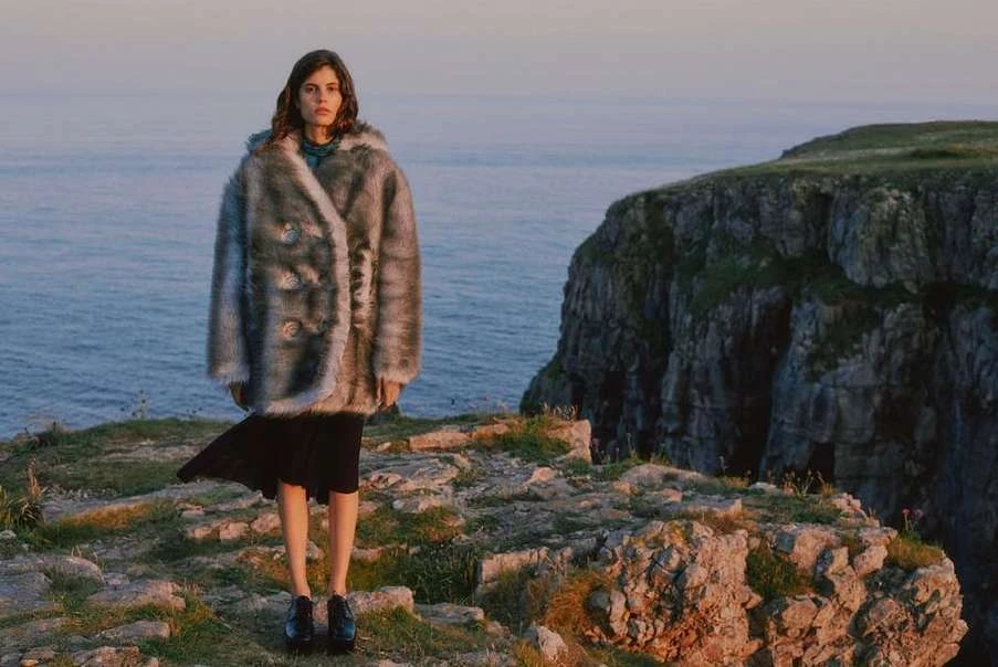 A woman stands on a cliff overlooking the ocean, wearing a furry coat and a dark skirt in a fashion editorial style photograph.