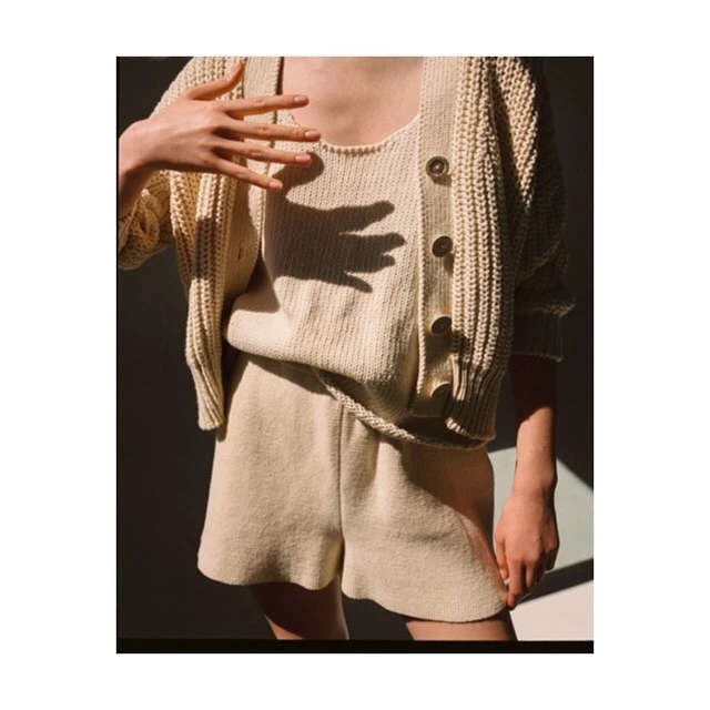 A person poses indoors wearing a matching knit shorts and top set with a cardigan, illuminated by shadows and soft light.