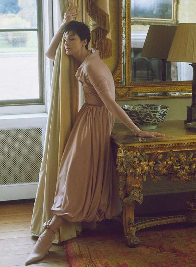 Woman with short dark hair in a pink dress posing beside a window and a gilded console table, captured by Agata Pospieszynska.
