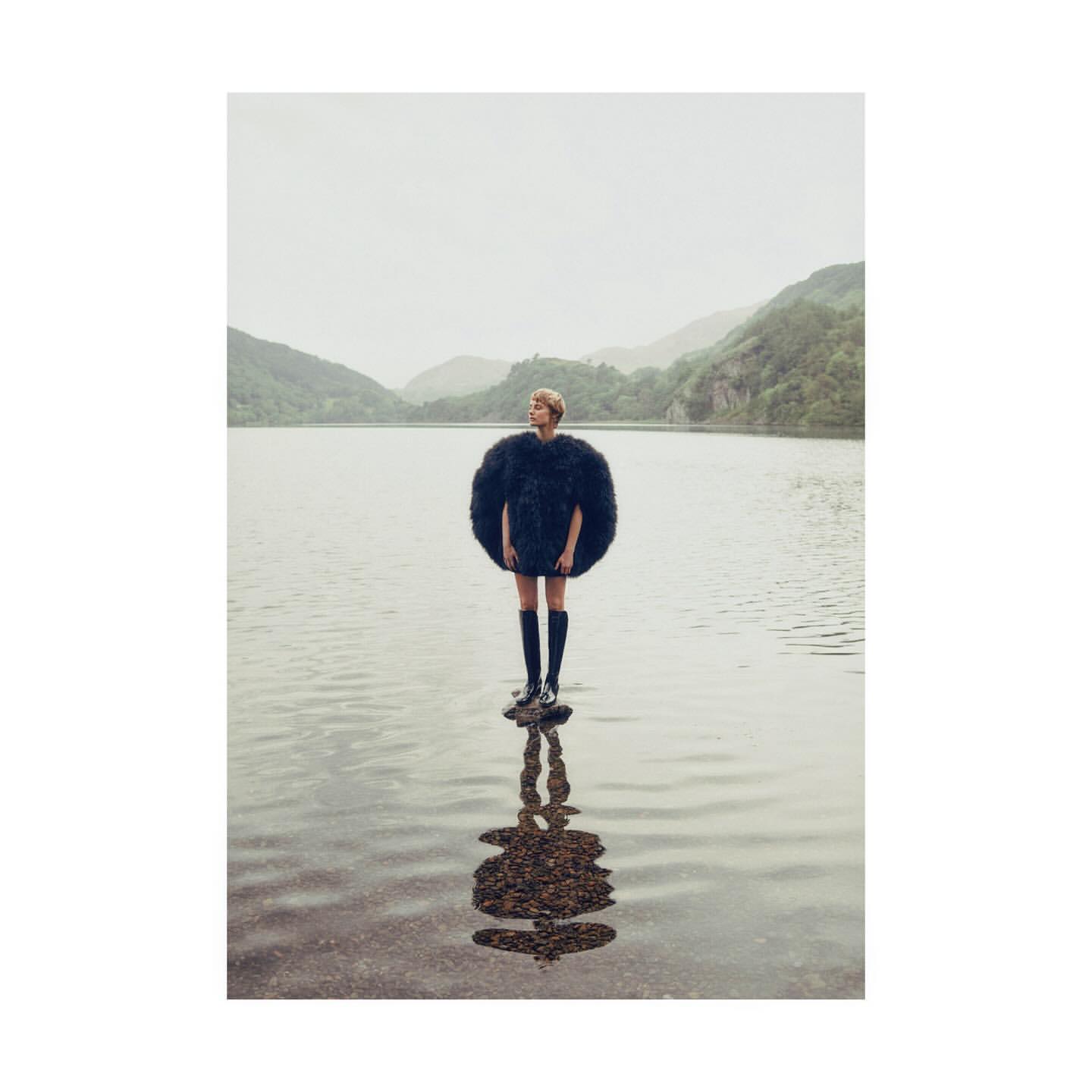 A woman stands on rocks in a lake wearing a large, round black fur coat with mountains in the background on a cloudy day.