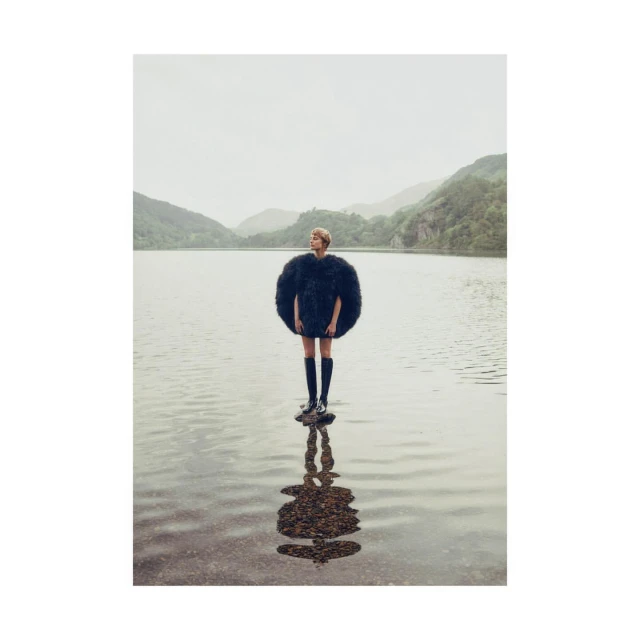 A woman stands on rocks in a lake wearing a large, round black fur coat with mountains in the background on a cloudy day.