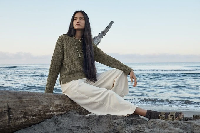 A woman sits on a log at the beach wearing a textured green sweater and beige pants, looking out at the ocean under a blue sky.