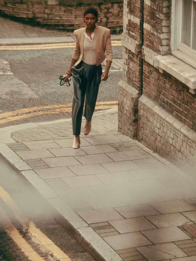 A woman in stylish beige jacket and dark pants walks on a city sidewalk, accented by brick buildings and street markings, captured outdoors.