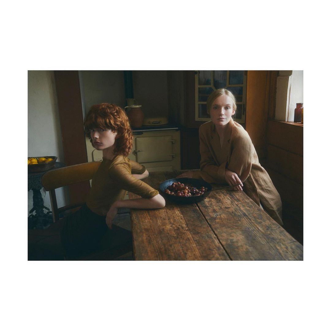 Two people sit at a rustic wooden table indoors with a bowl of fruit, bathed in soft natural light, creating a warm and inviting atmosphere.