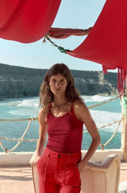 A woman with long brown hair poses in a red tank top and pants against a backdrop of the ocean, cliffs, and a partially visible white chair.