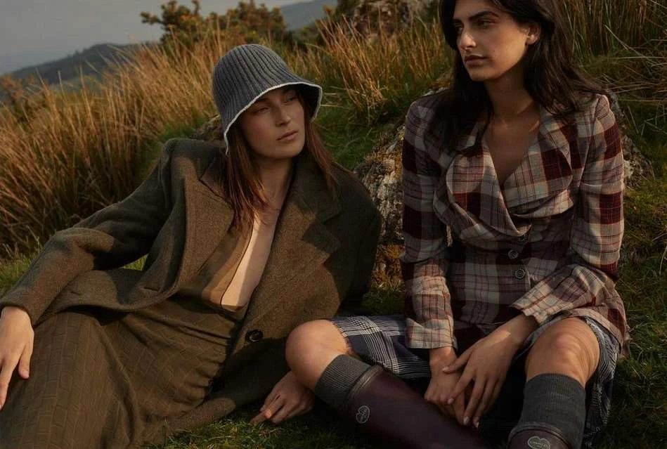 Two women in stylish fall clothing sitting on grass in a mountain field, one wearing a knit hat and coat, the other in a plaid jacket.